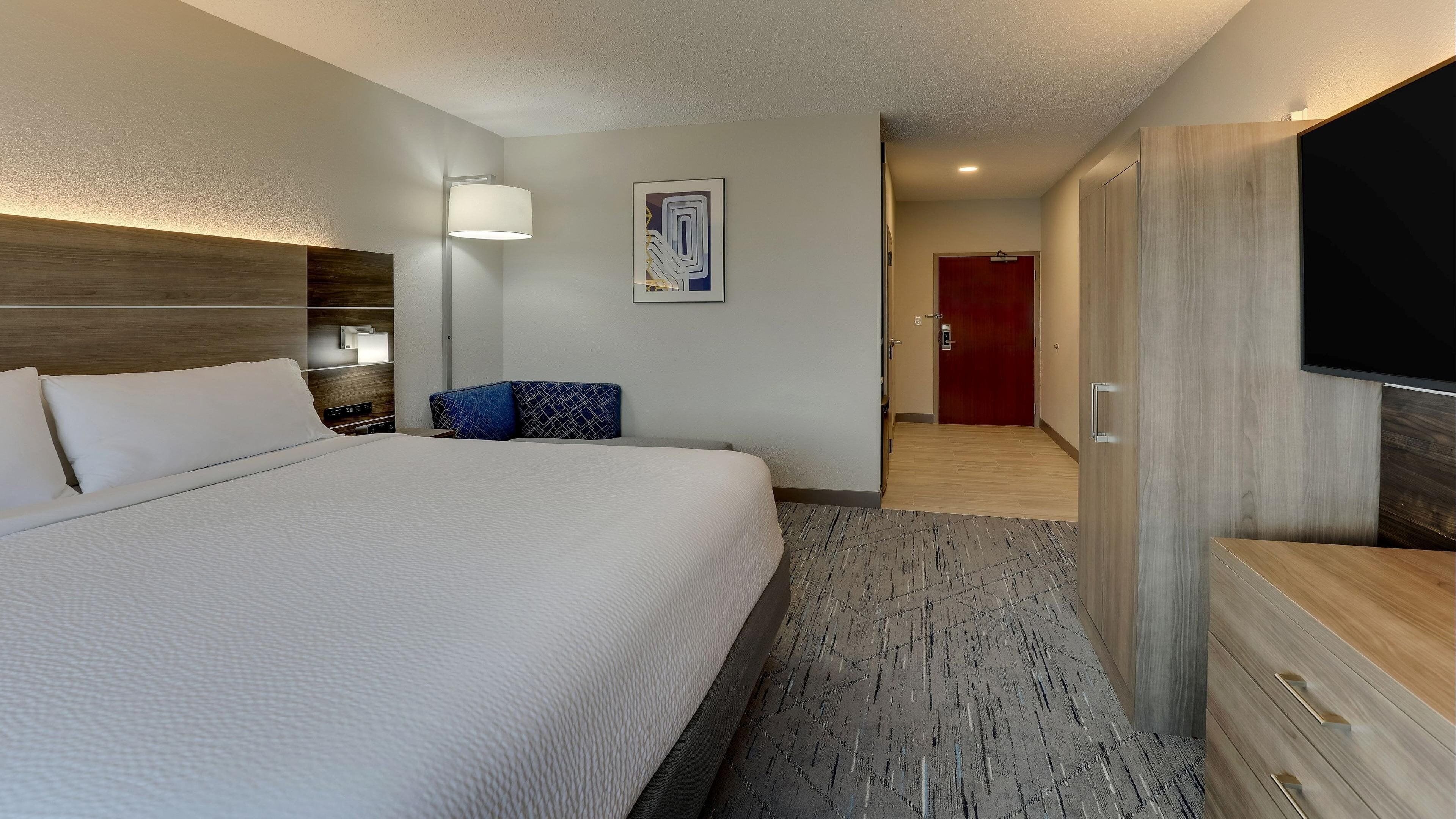 Una habitación de hotel con una cama grande, una mesita de noche con lámpara, un televisor, un armario y una puerta que da a otra habitación.