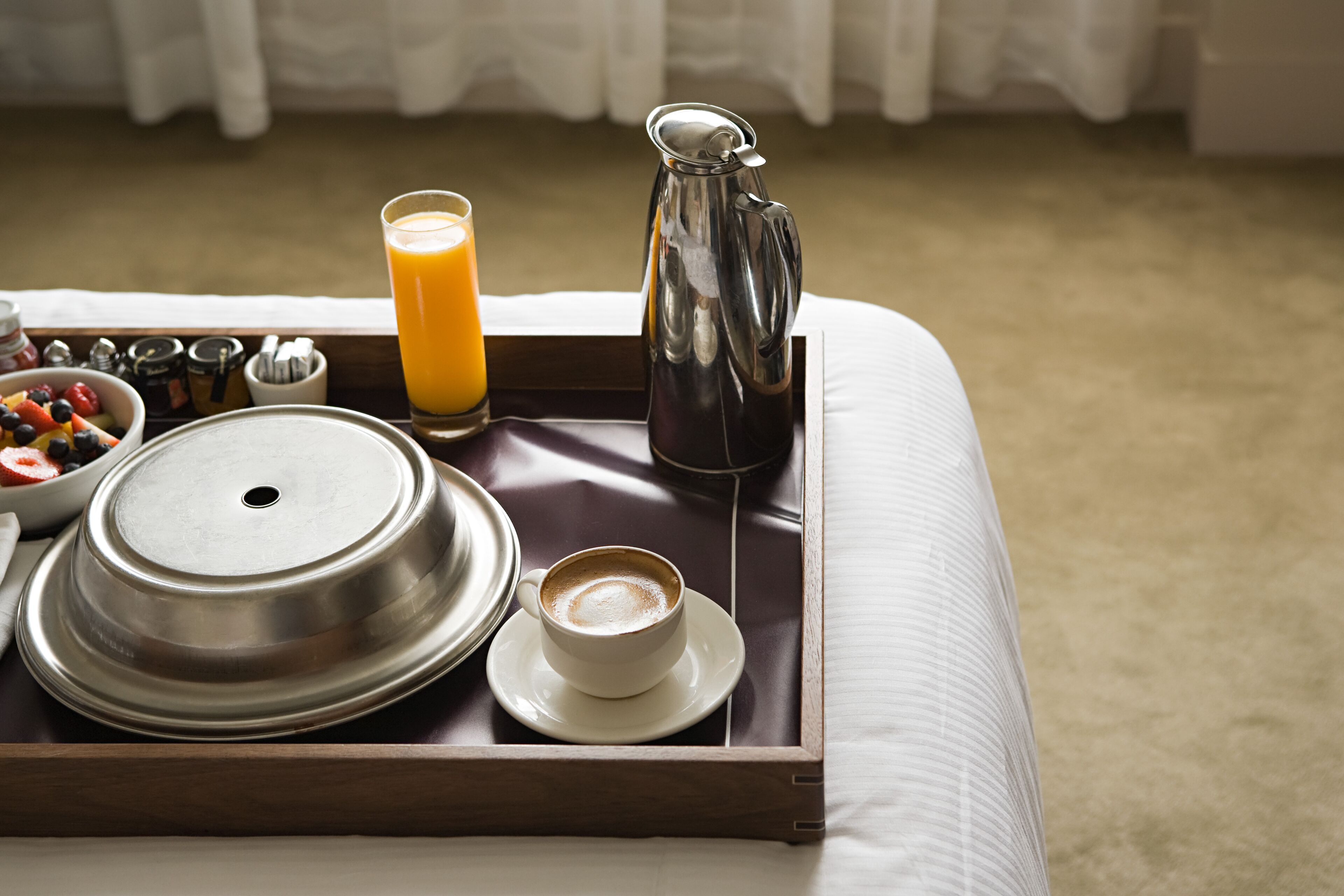 A breakfast tray with a coffee cup, a glass of orange juice, a pitcher, and a bowl of fruit on a bed.