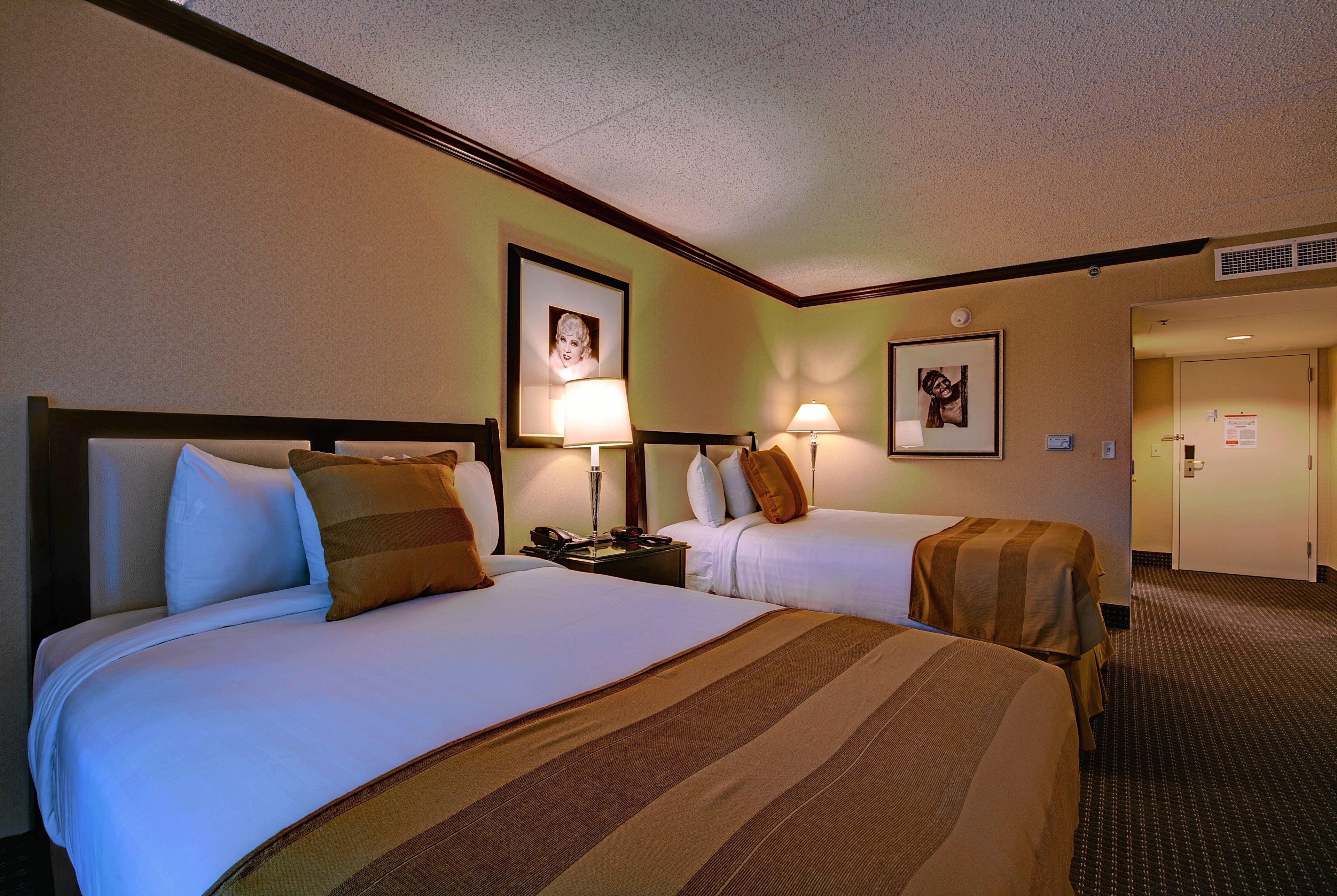 A hotel room with two beds, each with white linens and brown striped bedspreads. There are bedside lamps and framed pictures on the walls.