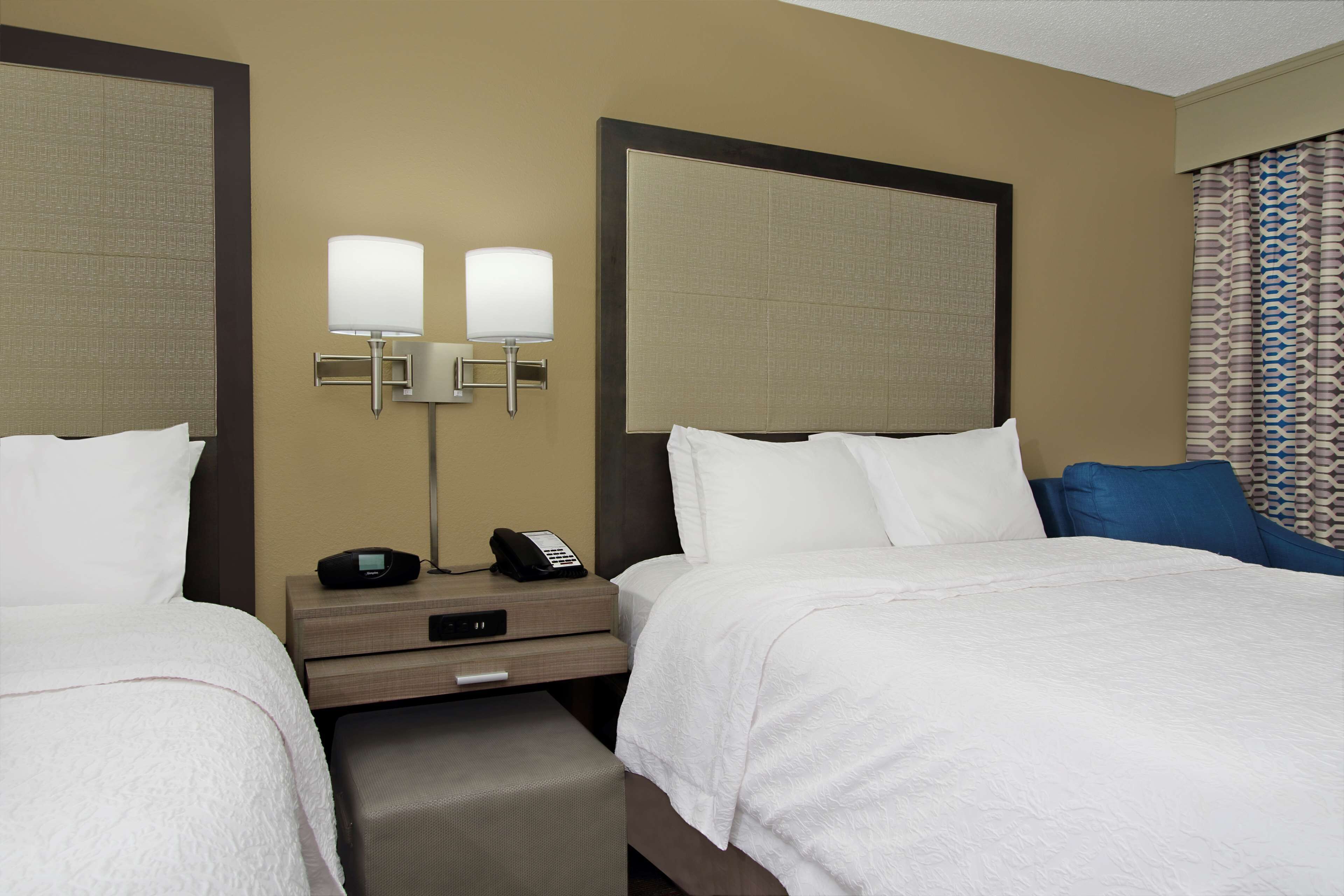 A hotel room with two beds, a bedside table with a phone and lamp, and a patterned curtain.