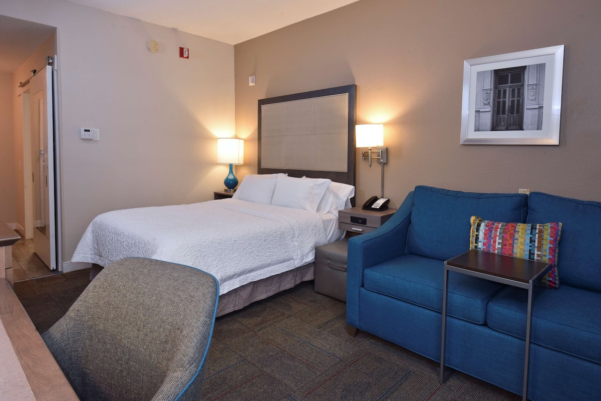 A hotel room with a bed, a blue sofa, a small table, and a nightstand with a lamp.
