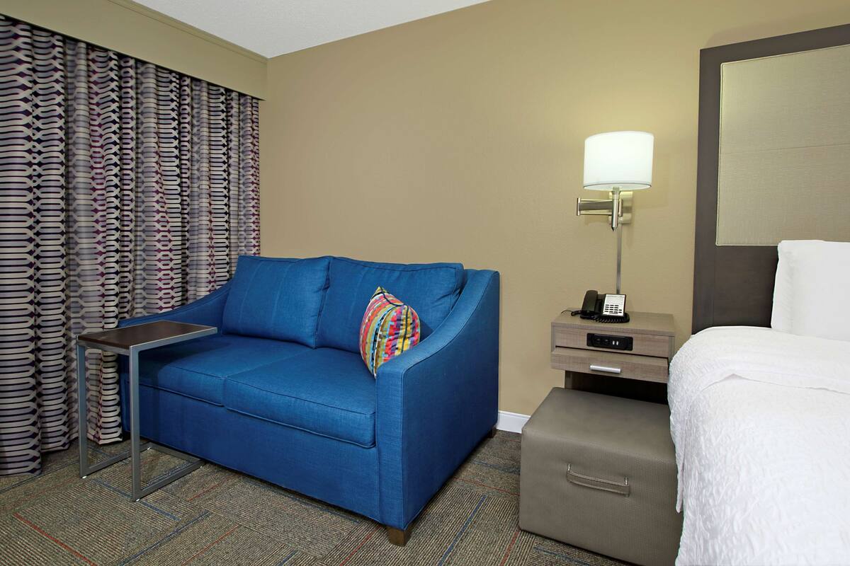 A hotel room with a blue sofa, a small side table, a lamp, a bed, and a suitcase.