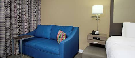 A hotel room with a blue sofa, a small side table, a lamp, a bed, and a suitcase.