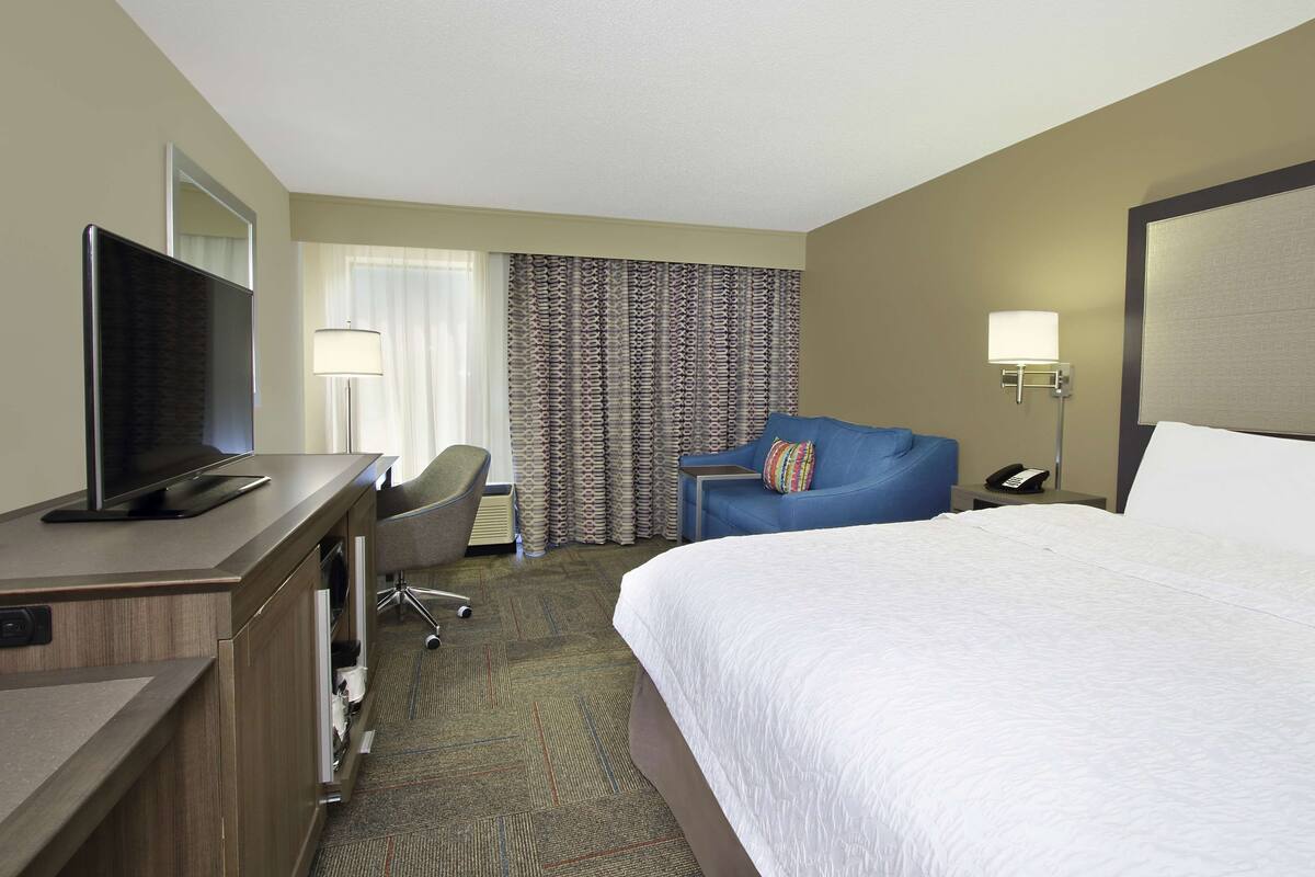 A hotel room with a bed, a desk with a TV, a chair, a lamp, and a phone on a side table.