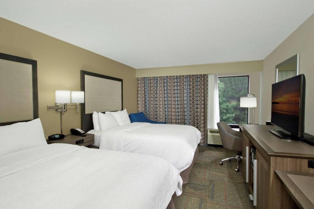 A hotel room with two beds, a desk, a chair, a TV, and a window with curtains.