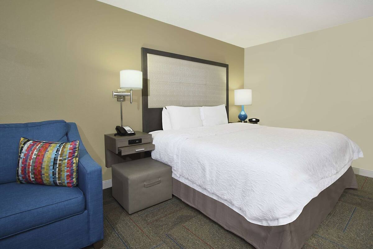 A hotel room with a bed, a desk, a chair, a nightstand, and a lamp.