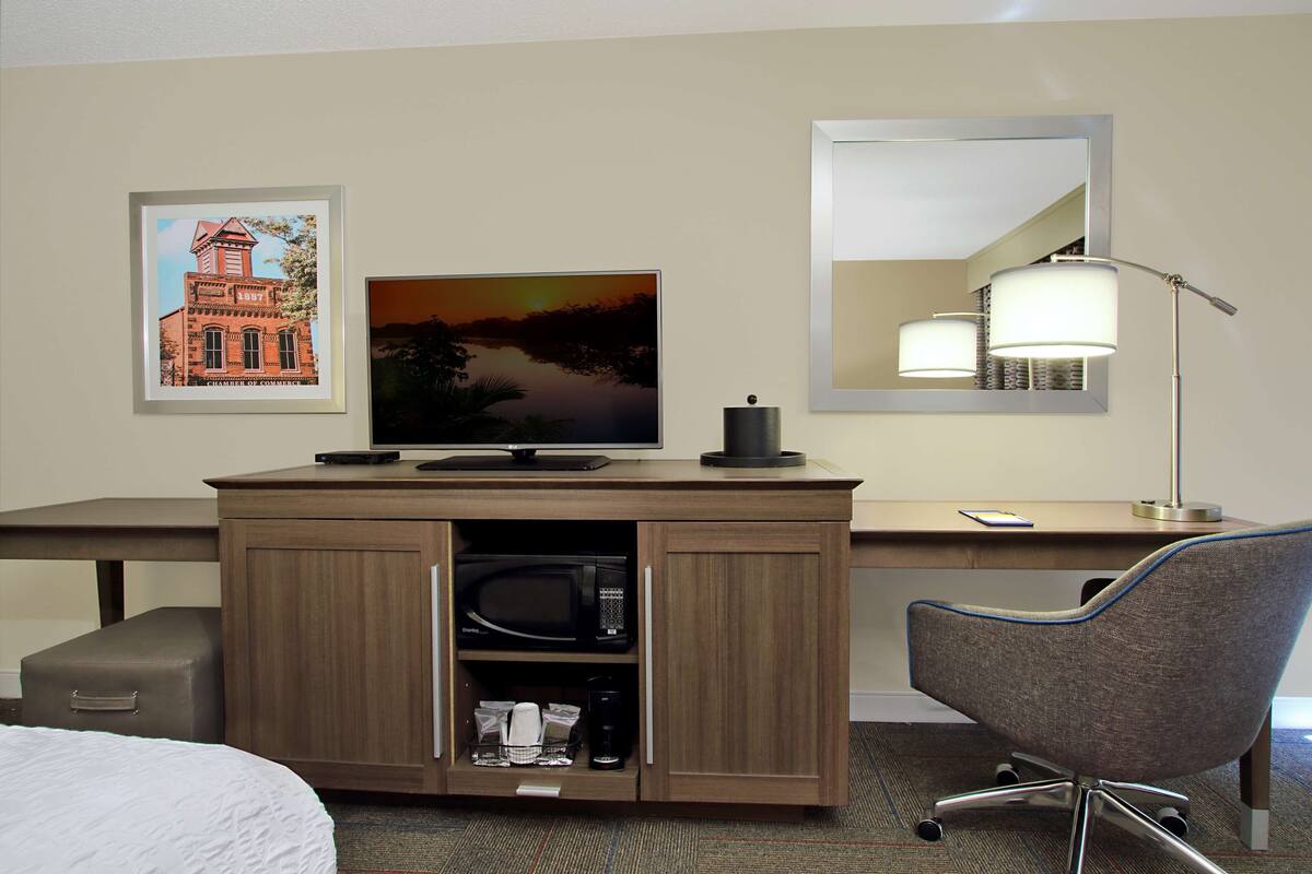 A hotel room with a wooden desk, a flat-screen TV, a microwave, a coffee maker, a chair, and a bed.