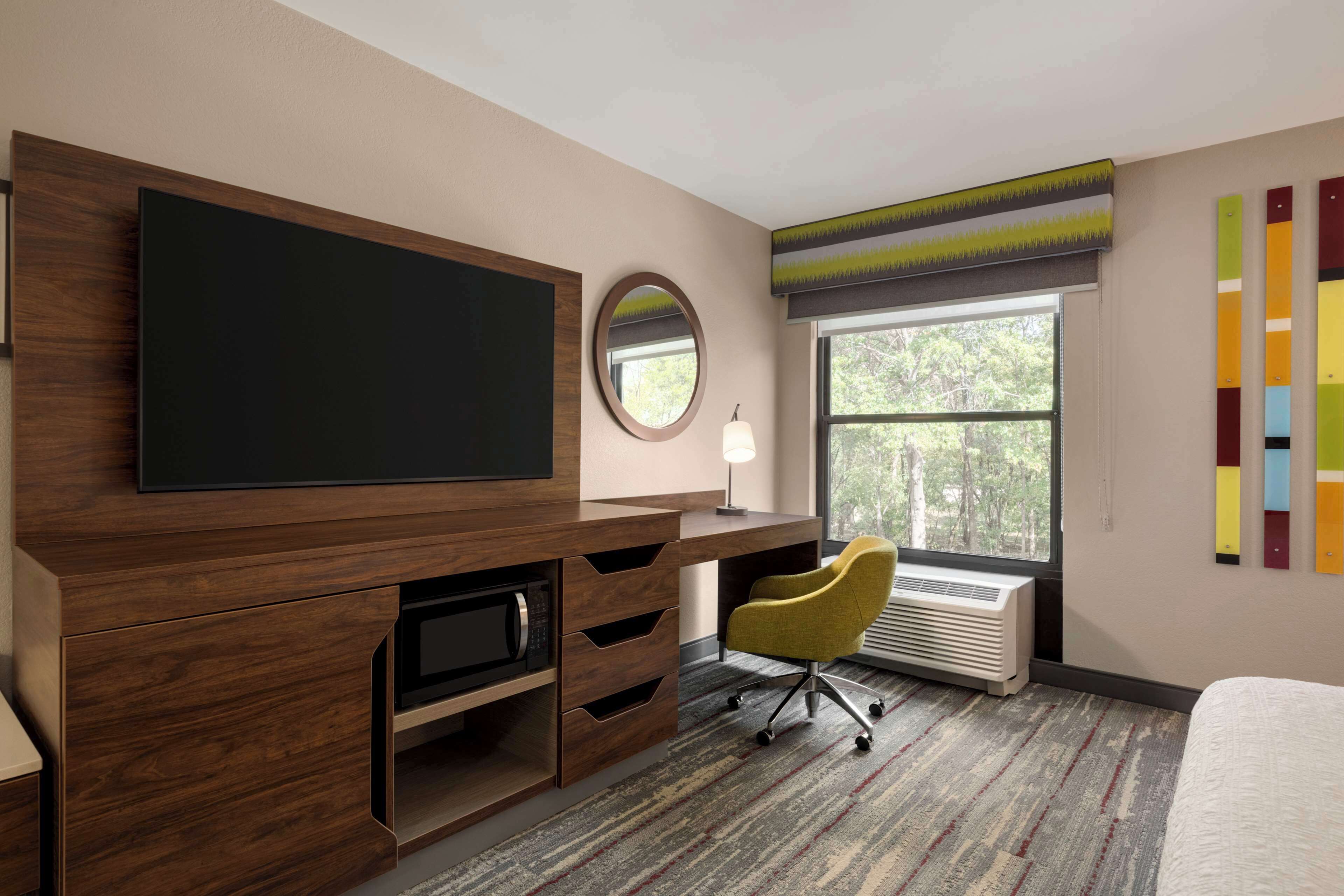 Quarto de hotel com uma televisão de tela plana grande, um espelho redondo, uma cadeira amarela, uma mesa com gavetas e uma janela com vista para árvores.