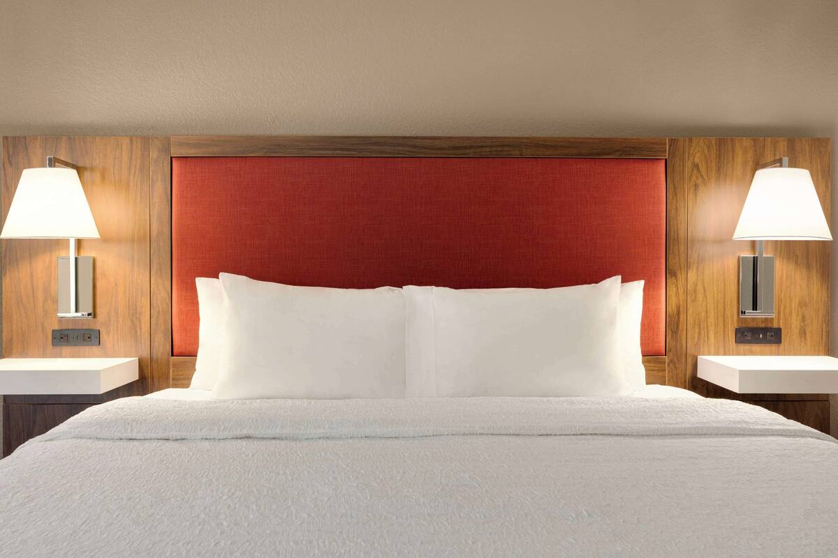 A hotel room with a large bed, two bedside tables with lamps, a wooden headboard, and a red accent wall.