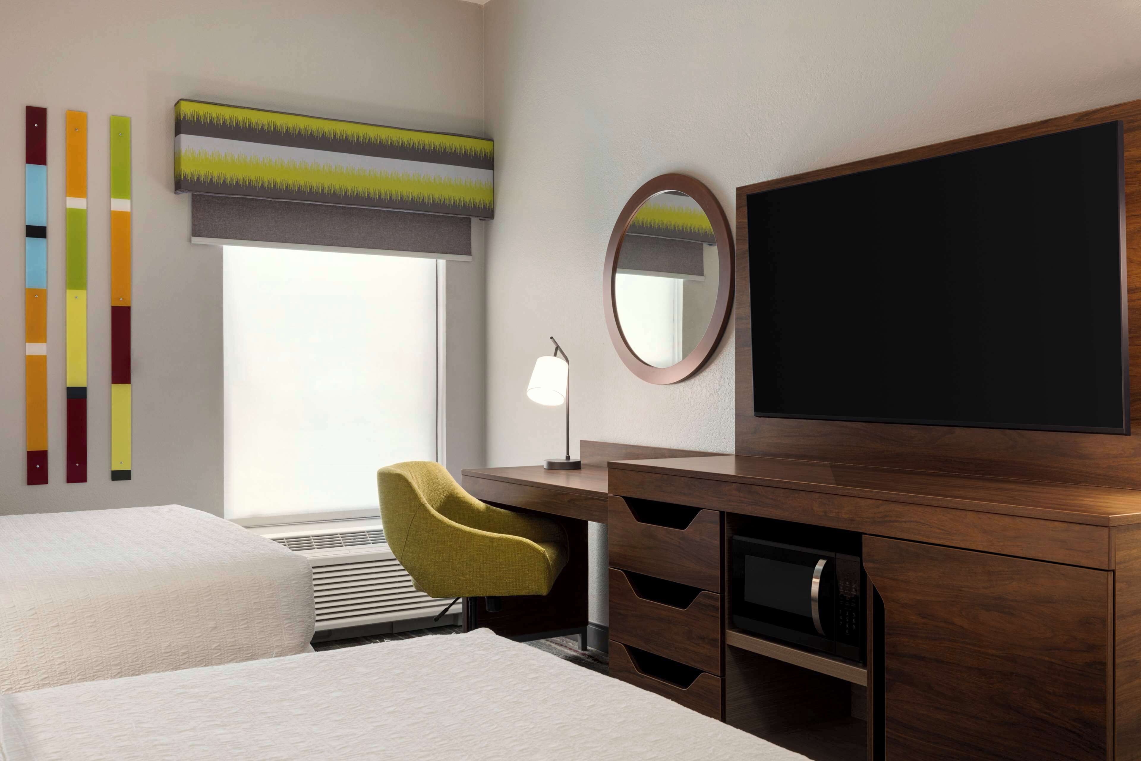 A hotel room with two beds, a desk with a chair, a television, a mirror, and a window with blinds.