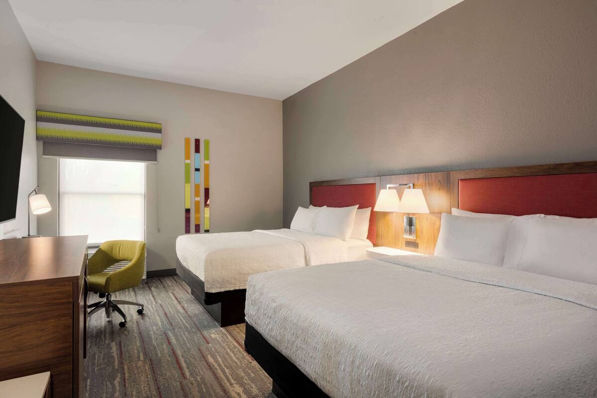 A hotel room with two beds, a desk, a chair, and a window with blinds.