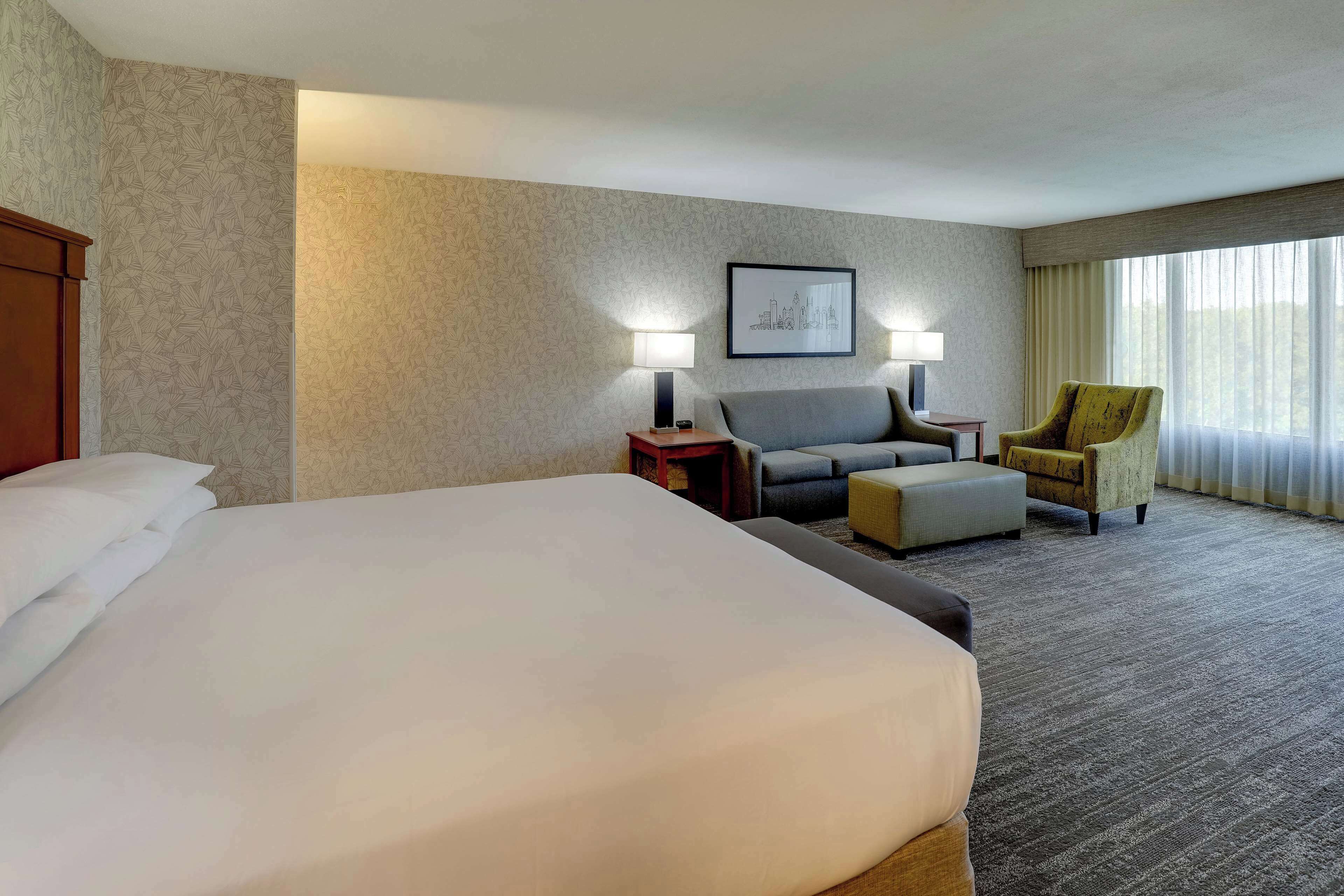 Une chambre d’hôtel avec un grand lit, un canapé, deux fauteuils, une petite table et un tableau représentant un paysage urbain, encadré et accroché au mur.