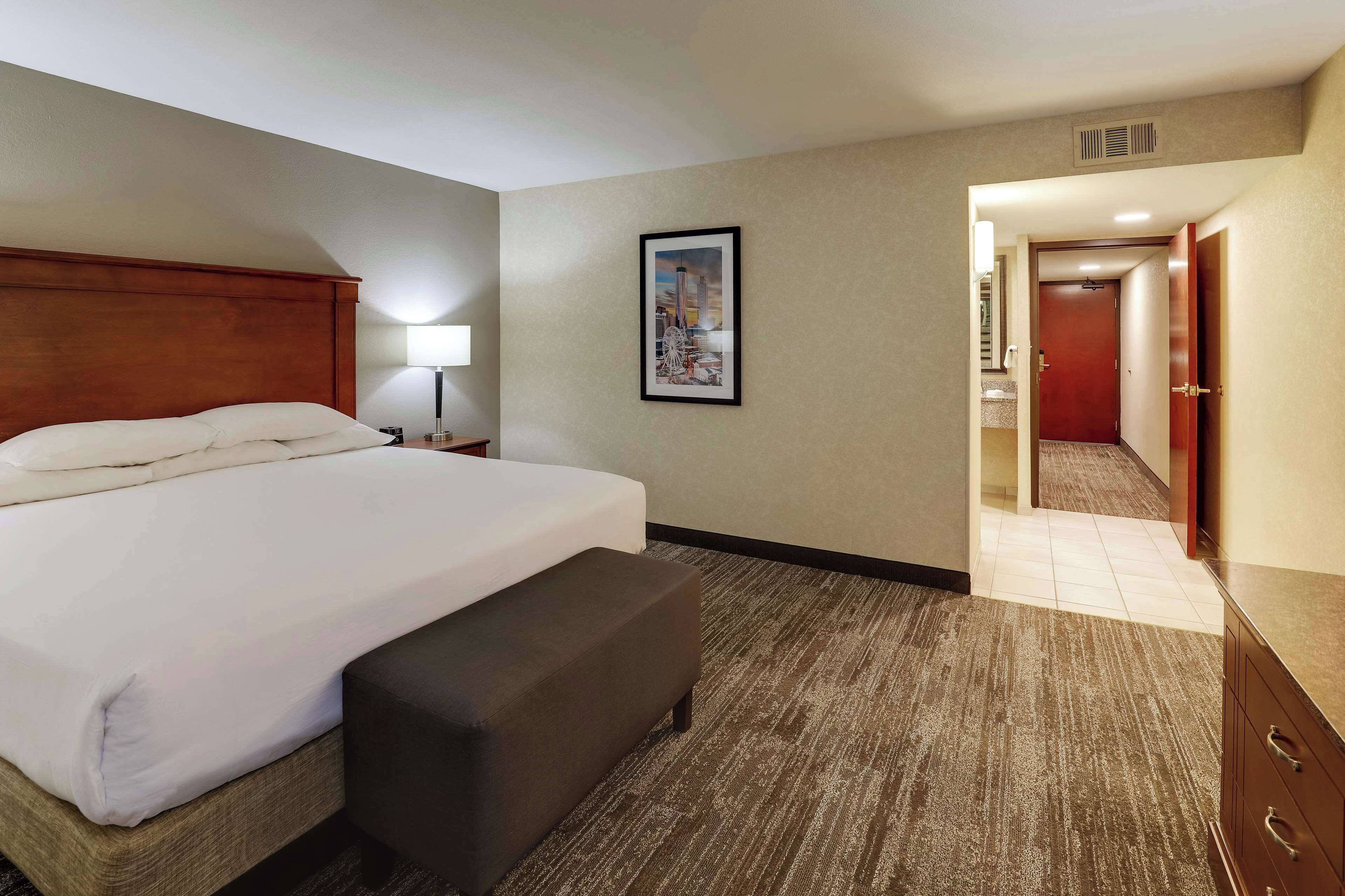 Una habitación de hotel con una cama grande, una lámpara de noche, un cuadro enmarcado en la pared y una puerta que da a un baño.