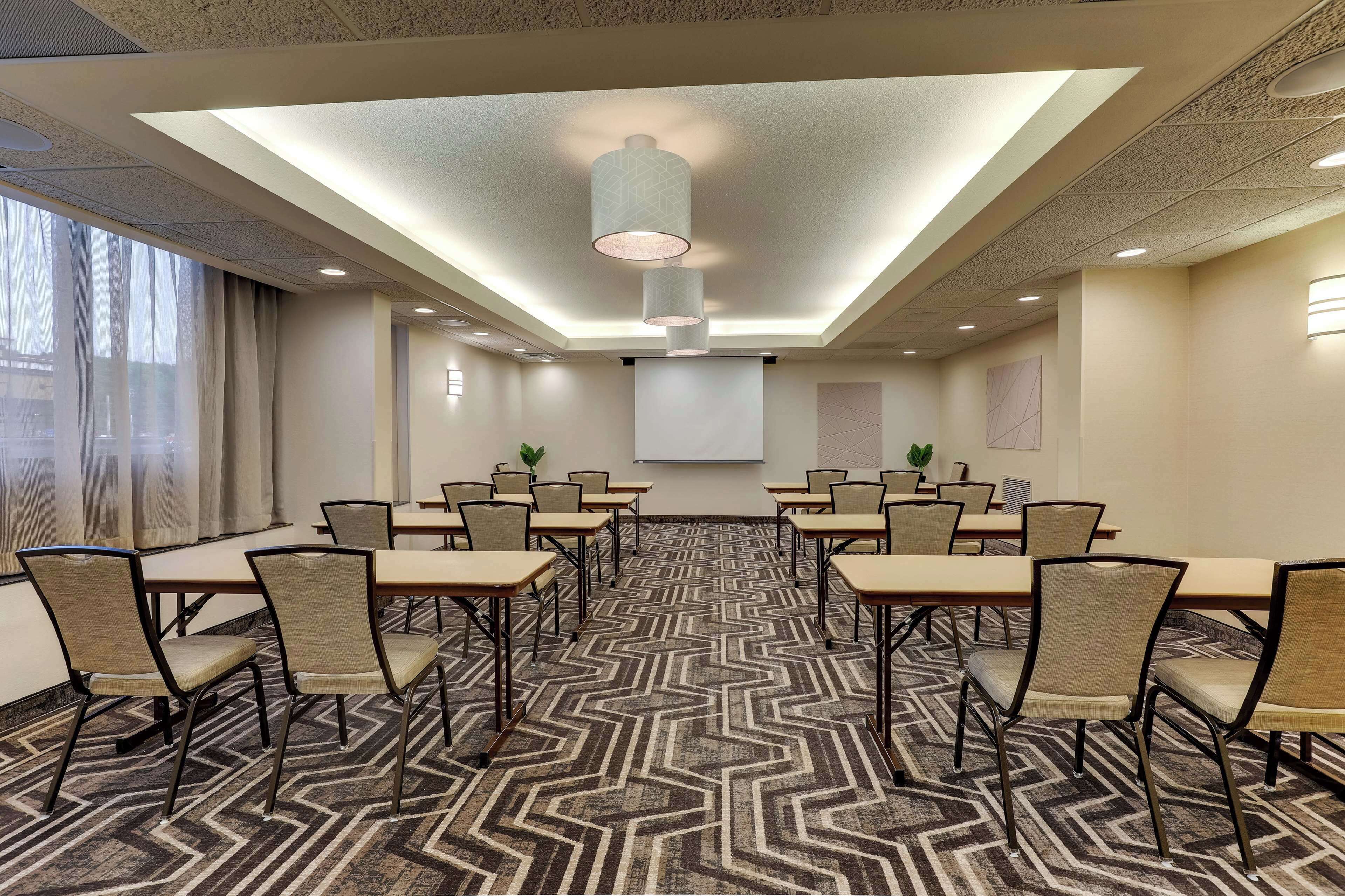 A conference room with a geometric patterned carpet, multiple tables with chairs, a projector screen, and a ceiling with recessed lighting.