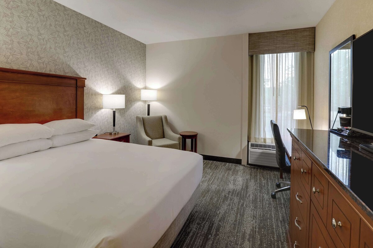 A hotel room with a large bed, a desk with a TV, a chair, and a lamp.