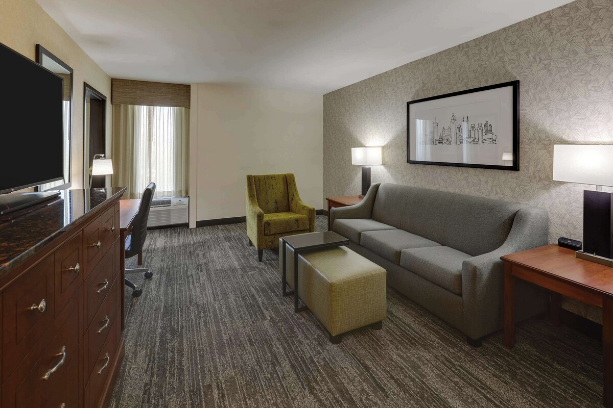 A hotel room with a TV, a sofa, a yellow armchair, a small table, and a lamp.