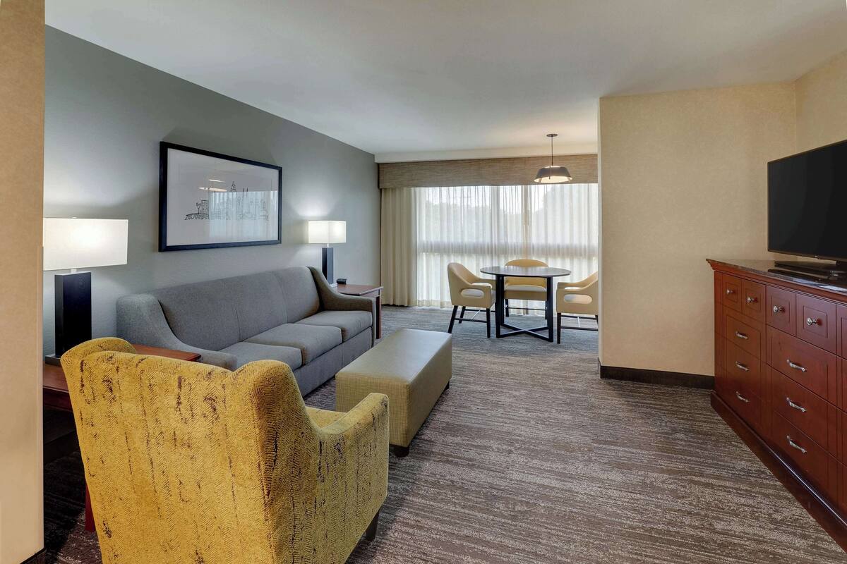 A hotel room with a grey sofa, yellow armchair, a small table with chairs, a TV, and a framed picture on the wall.