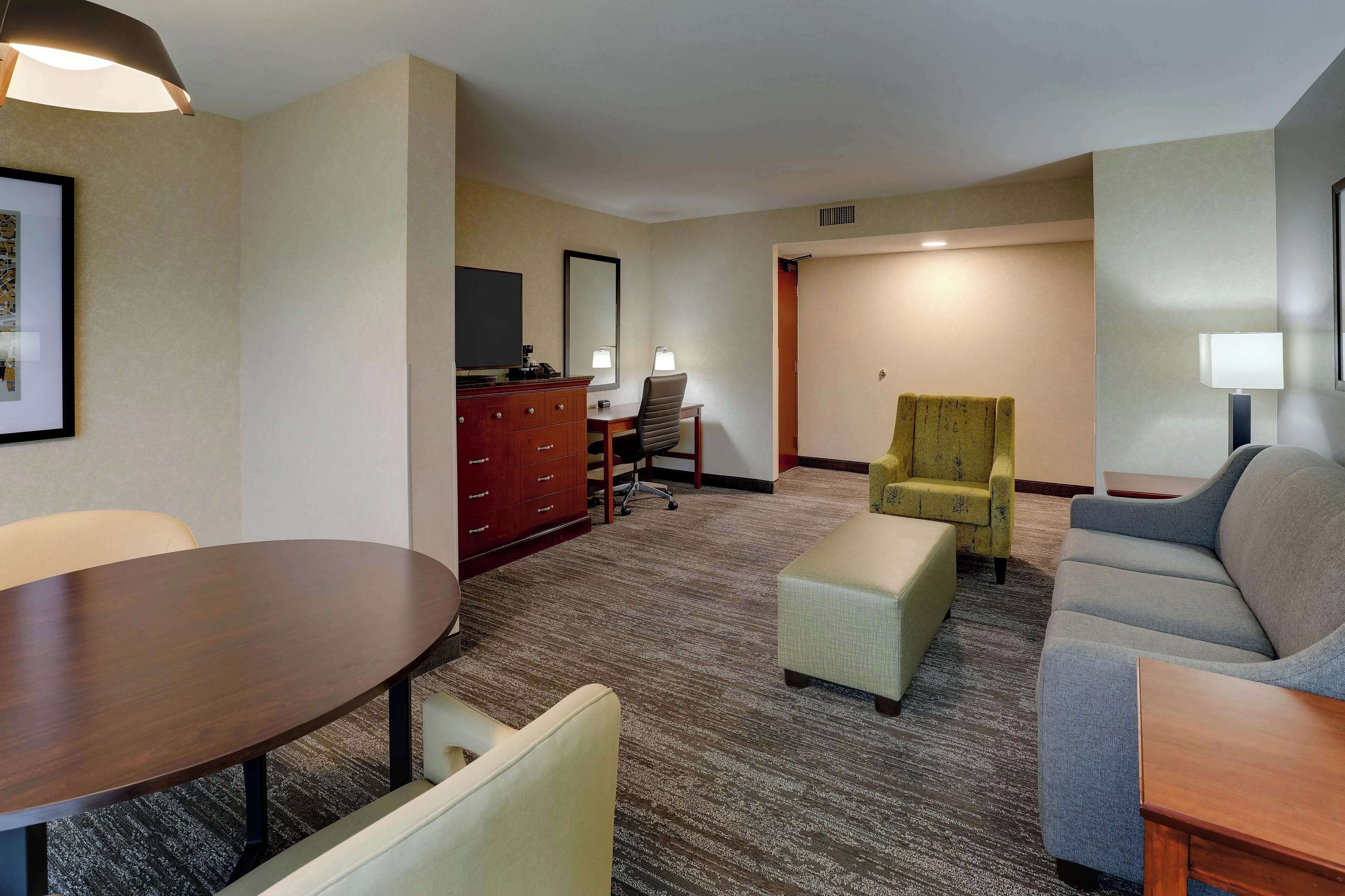 A hotel room with a dining table, a sofa, a small armchair, a desk with a chair, a TV, and a mirror.