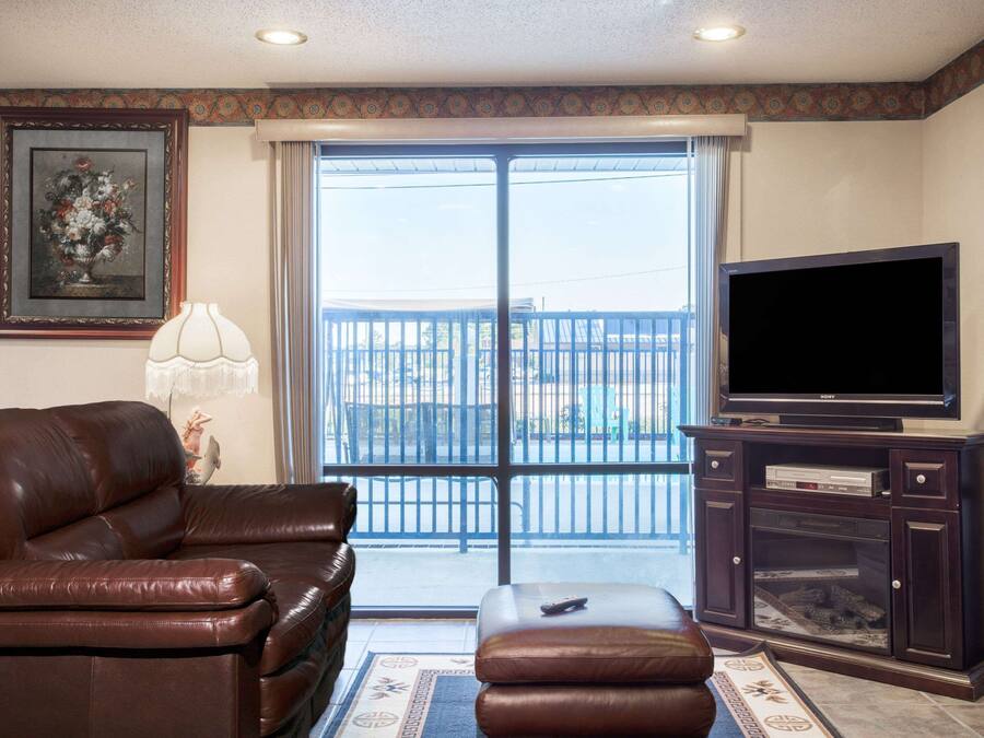 A living room with a brown leather sofa, a television on a wooden stand, and a large window with a view of a balcony.