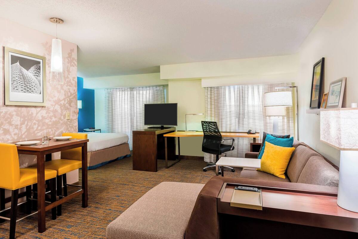 Ein Hotelzimmer mit Bett, Schreibtisch mit Computer, Fernseher, Sofa und Esstisch mit Stühlen.