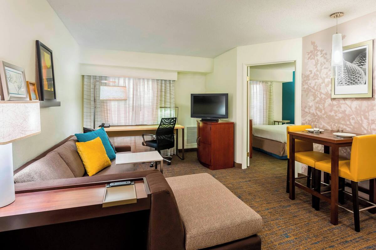 Ein Hotelzimmer mit einer Couch, einem kleinen Esstisch mit gelben Stühlen, einem Fernseher und einem Schreibtisch mit Stuhl.