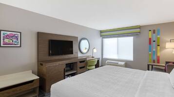 Habitación de hotel con cama, televisión, escritorio, silla, ventana con vistas y una obra de arte colorida en la pared.
