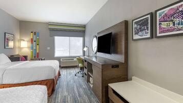 Ett hotellrum med två sängar, ett skrivbord med en platt-TV, en skrivbordslampa, en stol, en bänk och ett fönster med utsikt.