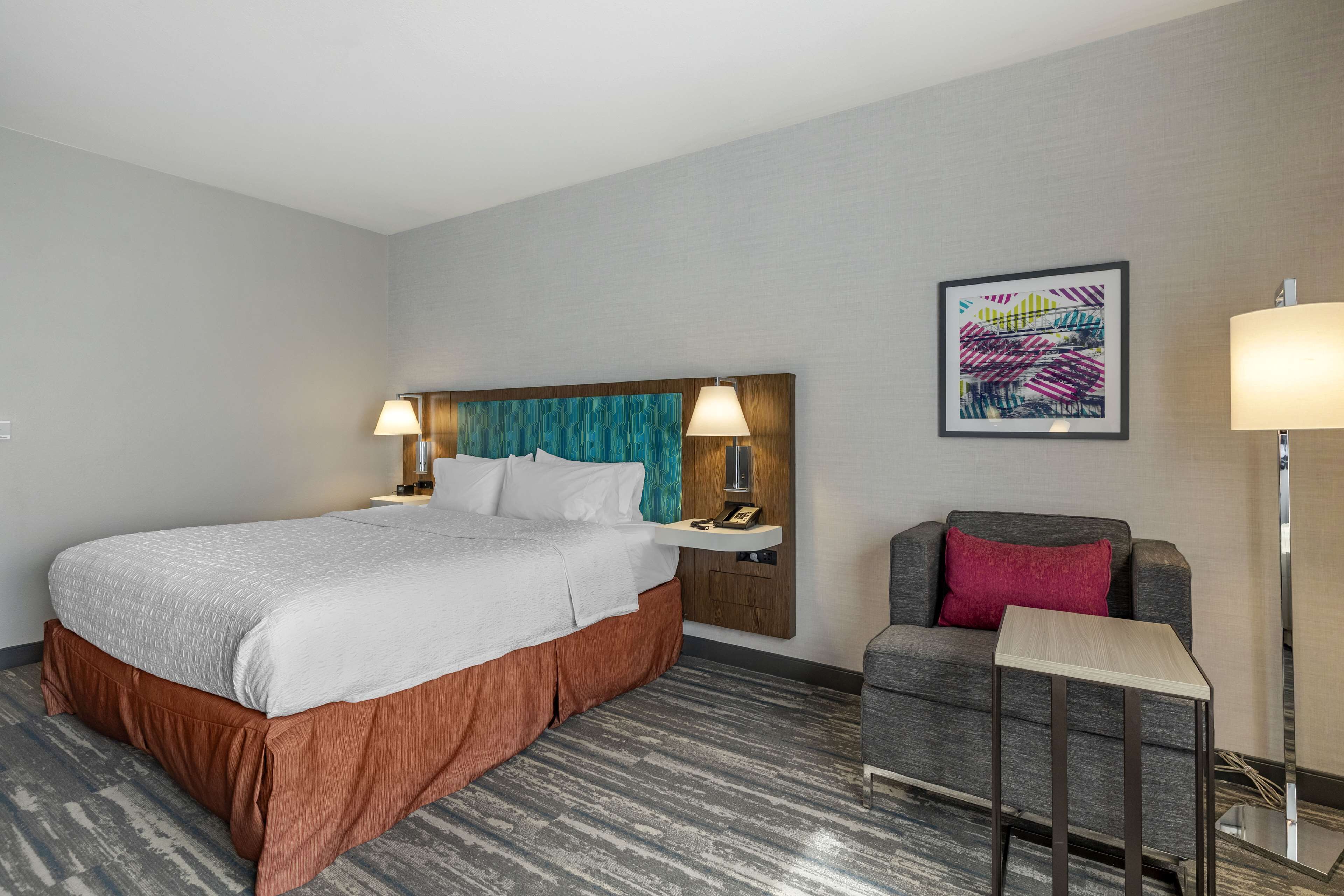 Ein Hotelzimmer mit Bett, Nachttisch, Stuhl, Lampe und einem Wandbild.