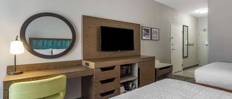 Habitación de hotel con cama, escritorio con silla, televisor, espejo y puerta que da al baño.