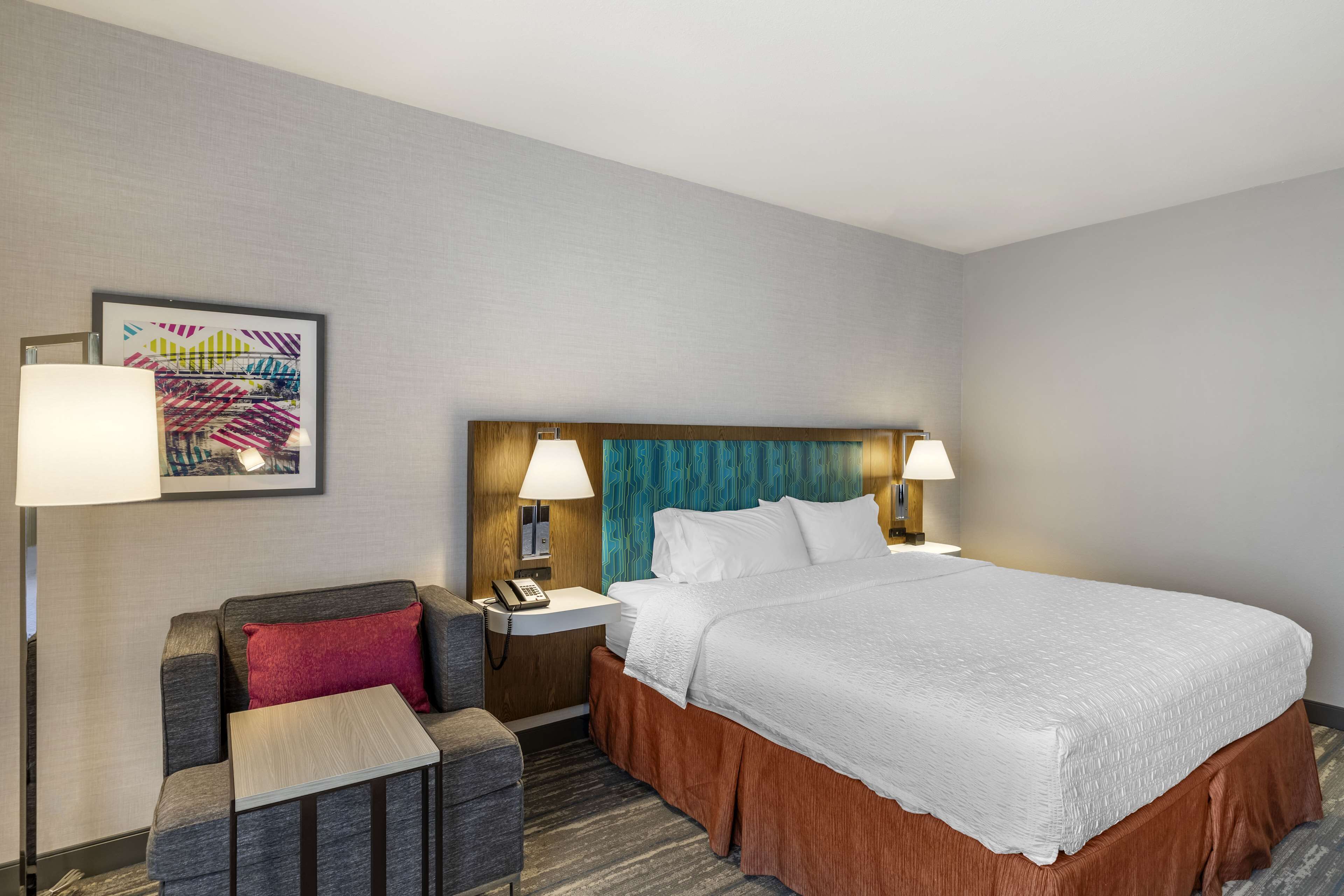 Une chambre d’hôtel avec un grand lit, un bureau avec une lampe, une chaise et une œuvre d’art colorée accrochée au mur.