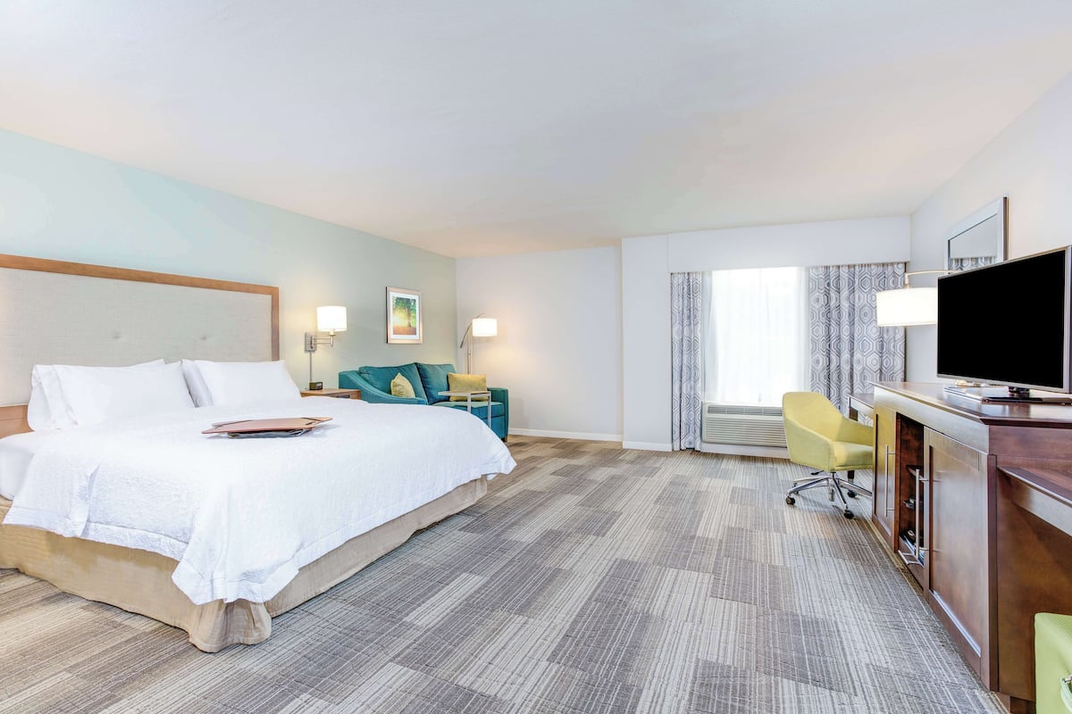Ein Hotelzimmer mit einem großen Bett, einem Schreibtisch mit Flachbildfernseher, einem Sessel und einer Schreibtischlampe.
