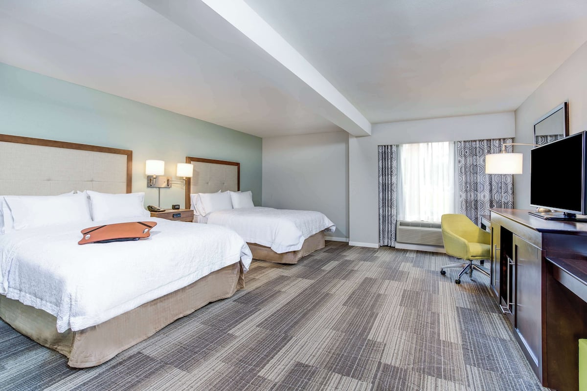 Ein Hotelzimmer mit zwei Betten, einem Schreibtisch mit Fernseher, einem Sessel und einem Fenster mit Vorhängen.