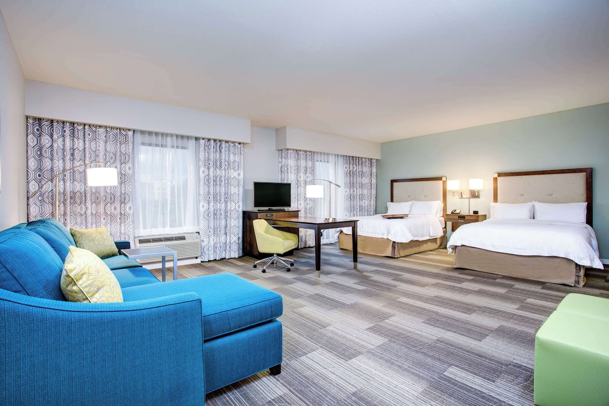 Ein Hotelzimmer mit blauem Sofa, einem grünen Hocker, zwei Einzelbetten, einem Schreibtisch mit Stuhl, einem Fernseher und einem Fenster mit gemusterten Vorhängen.