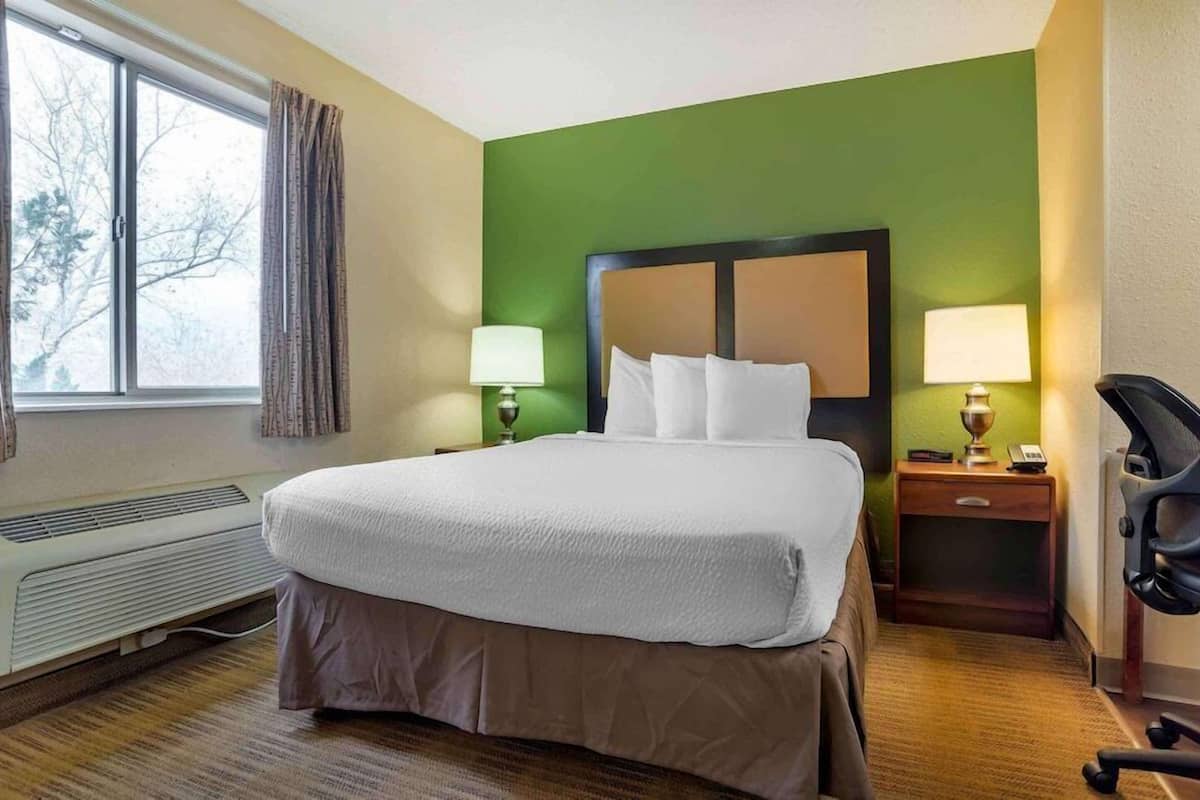 A hotel room with a large bed, a bedside table with a lamp, a chair, and a window with curtains.