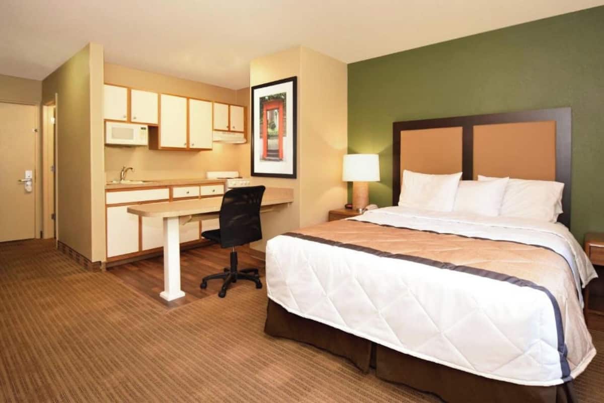 A hotel room with a bed, desk, chair, and a microwave in the kitchenette.