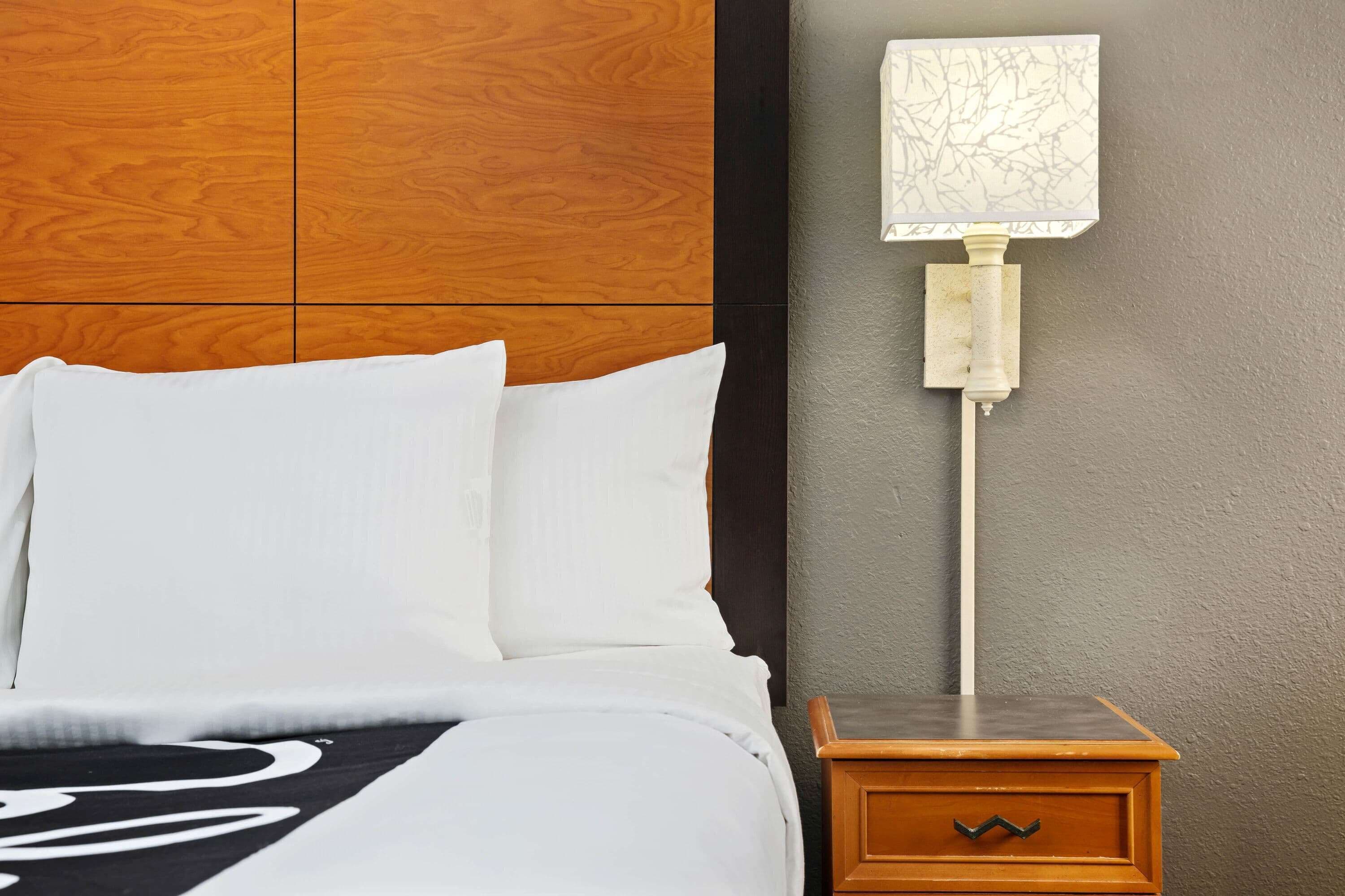A neatly made bed with white linens and a black and white blanket, a wooden nightstand with a lamp, and a textured wall behind.