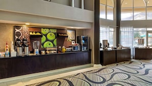 A hotel lobby with a counter featuring a 'La Quinta' sign, a coffee machine, a microwave, and a fruit display. - La Quinta Inn & Suites by Wyndham Macon (Macon)