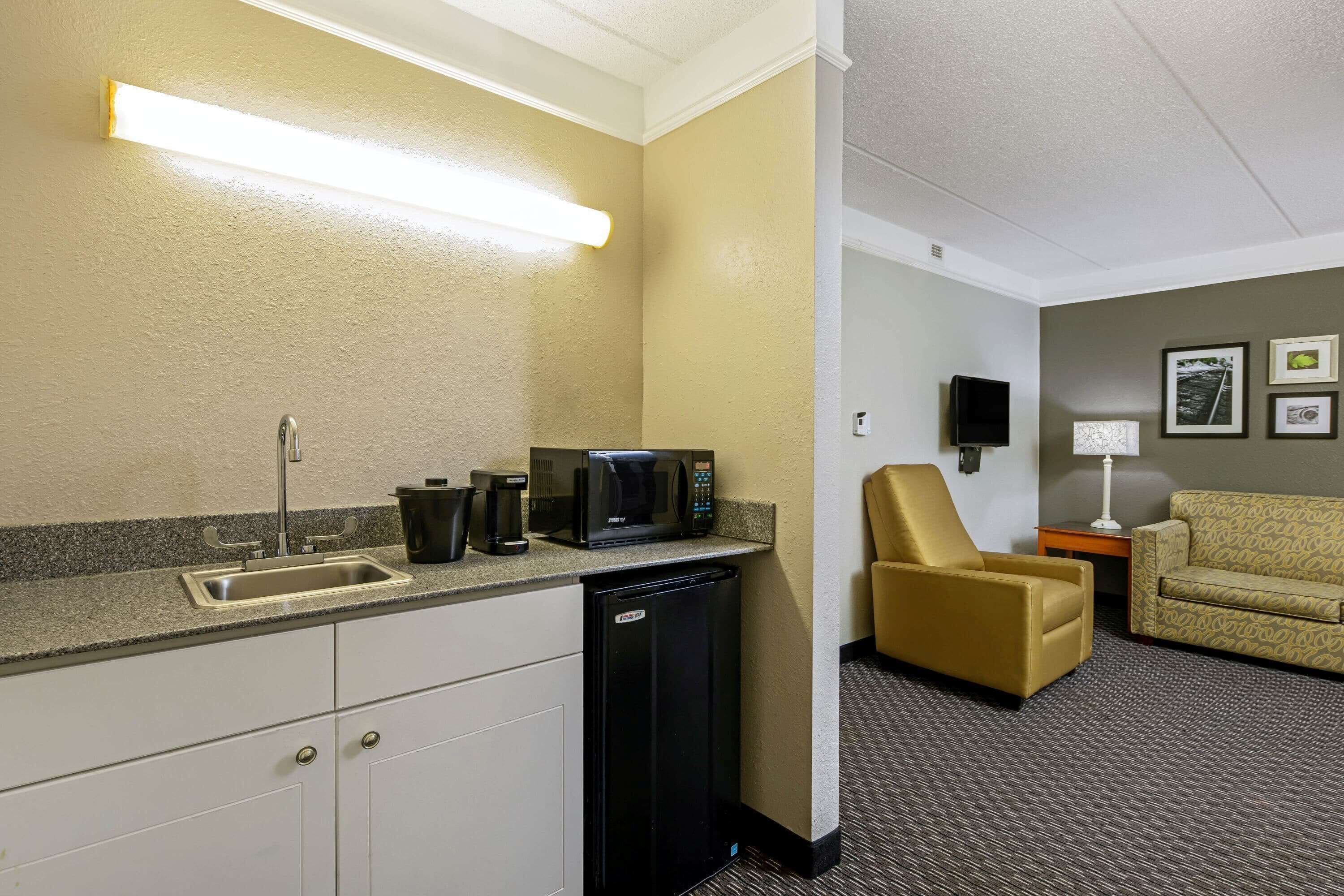 A compact hotel room with a kitchenette, a sofa, a chair, and a small table with a lamp.
