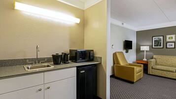 A compact hotel room with a kitchenette, a sofa, a chair, and a small table with a lamp.