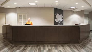 Un área de recepción con un mostrador de madera oscura, una persona con una camisa naranja detrás y una gran decoración de flor de loto montada en la pared.