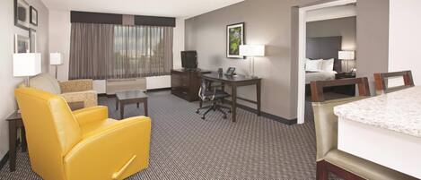 Habitación de hotel con un sillón amarillo, un escritorio, una mesa de comedor y una cama con cabecera.