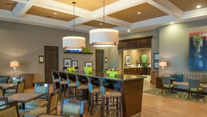 A hotel lobby with a bar area, dining tables, and chairs. There are hanging lamps, a reception desk, and a painting on the wall.