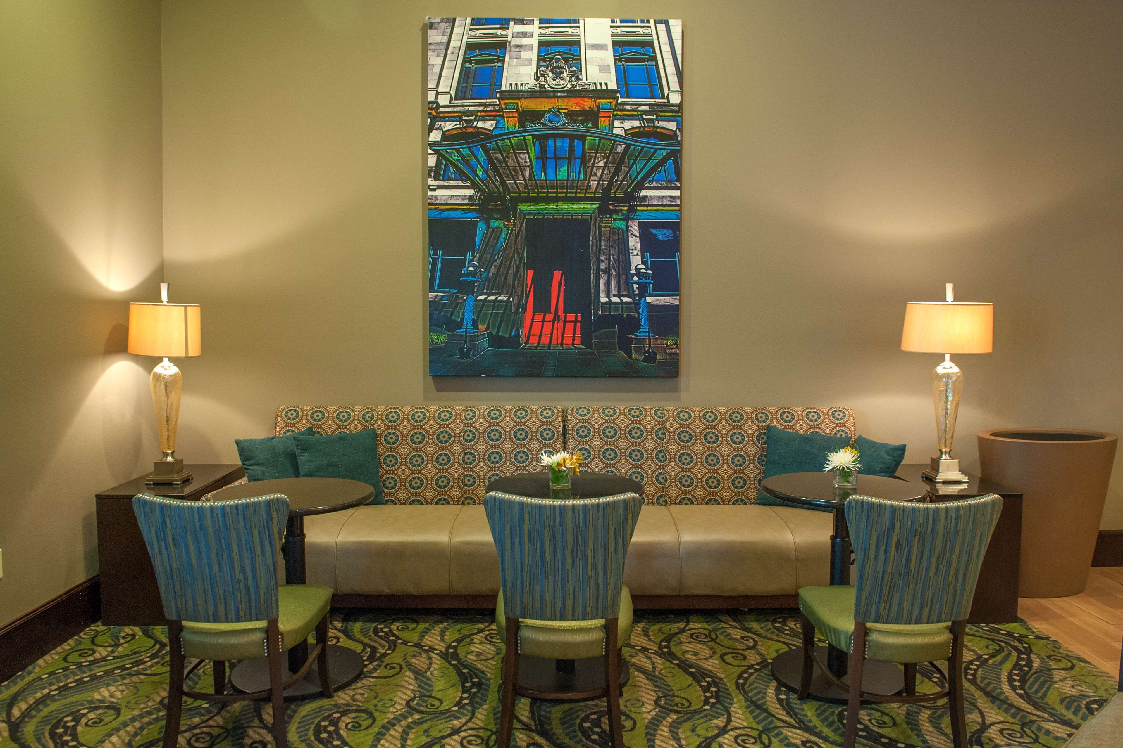 Quarto de hotel com um sofá estampado, duas cadeiras, uma mesa e um quadro de um edifício.