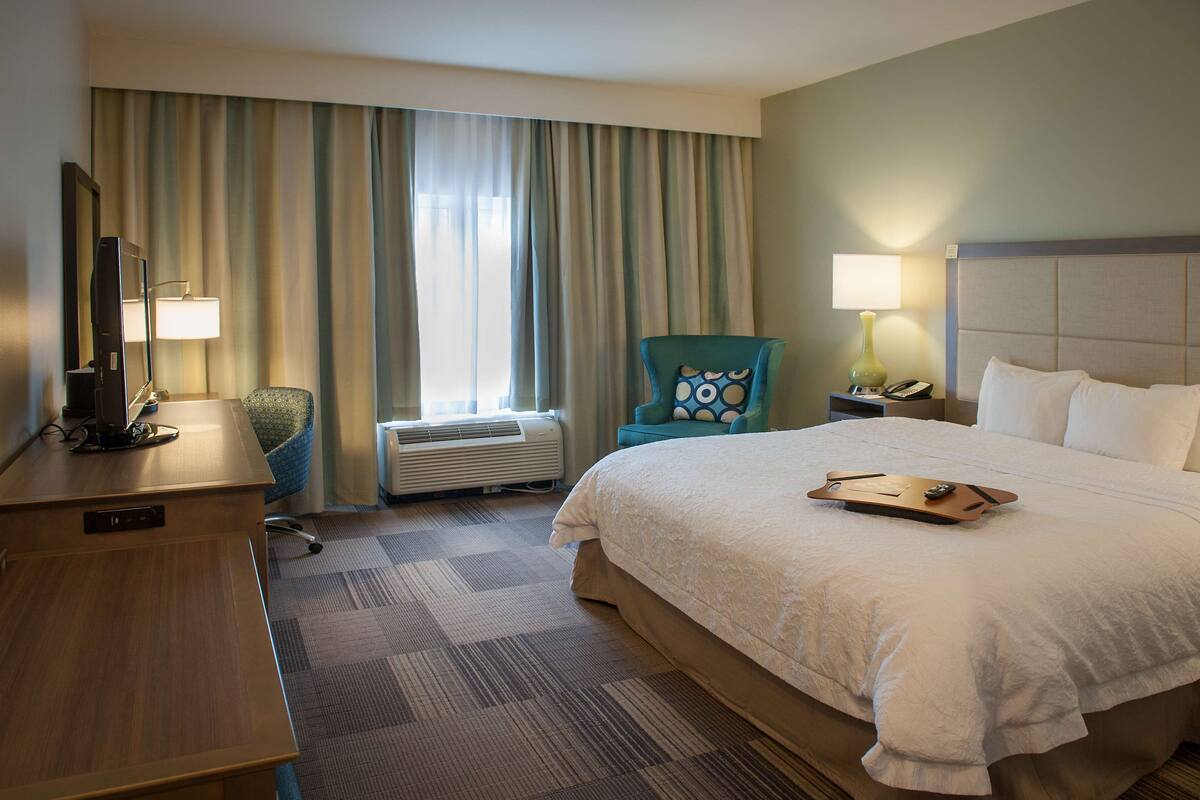 A hotel room with a large bed, a desk, a chair, a desk lamp, a bedside table with a phone, and a window with curtains.