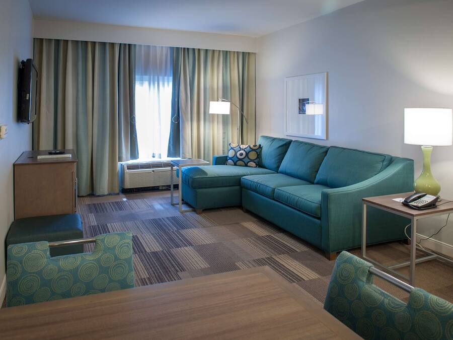 A hotel room with a teal sofa, a small table, a lamp, a TV mounted on the wall, and a window with curtains.