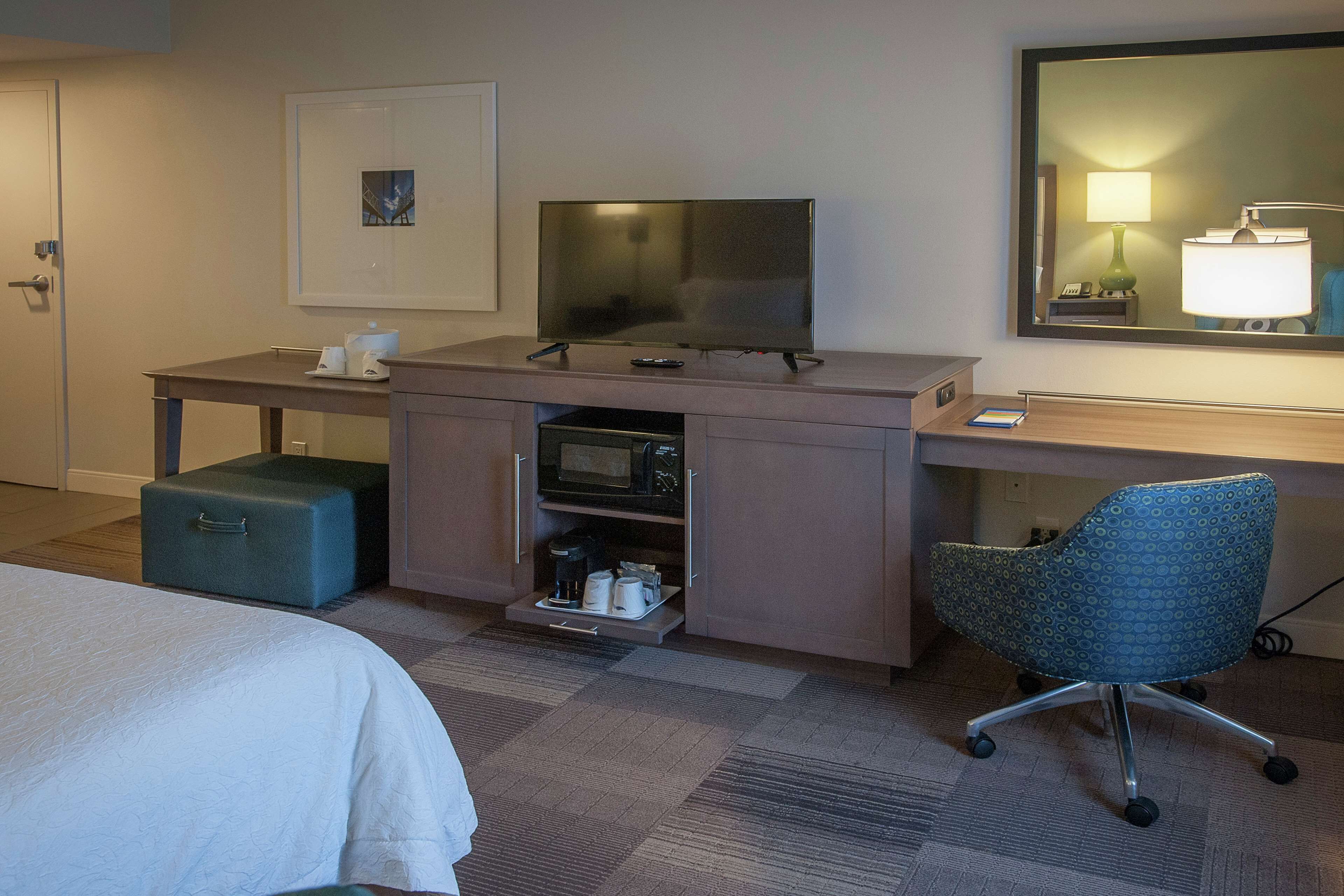 Habitación de hotel con cama, escritorio con televisor, microondas, cafetera, silla y pequeña mesa con lámpara.