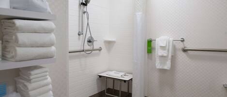 A bathroom with white tiled walls, a shower with a handheld showerhead, a toilet, and a small table with a towel.