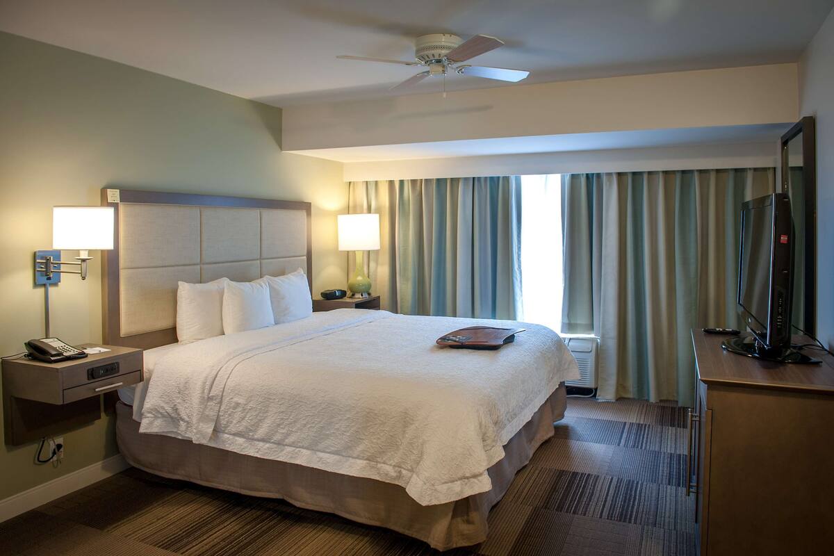 A hotel room with a large bed, a desk with a computer, a ceiling fan, and a window with curtains.