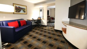 Ein Hotelzimmer mit blauem Sofa, roten Kissen, Schreibtisch, Stuhl, Flachbildfernseher und einem gemusterten Teppich.