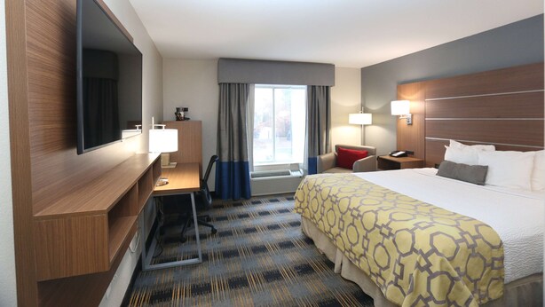 Ein Hotelzimmer mit einem großen Bett, einem Schreibtisch, einem Flachbildfernseher, einem Fenster mit Vorhängen und einem Nachttisch mit einer Lampe.